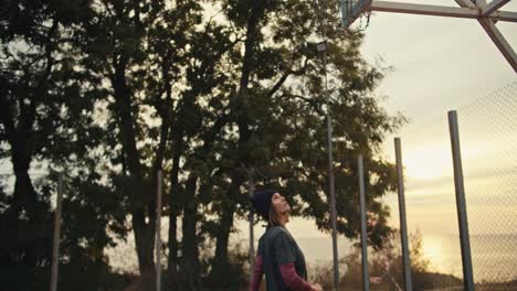 A-blonde-girl-with-a-bob-hairstyle-in-a-black-hat-runs-to-a-basketball-hoop-and-throws-the-ball-on-the-red-court-early-in-the-morning-during-her-training-in-the-summer