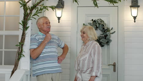 Senior-couple-together-in-porch-at-home.-Mature-man-stands-and-his-wife-comes-to-embrace-him