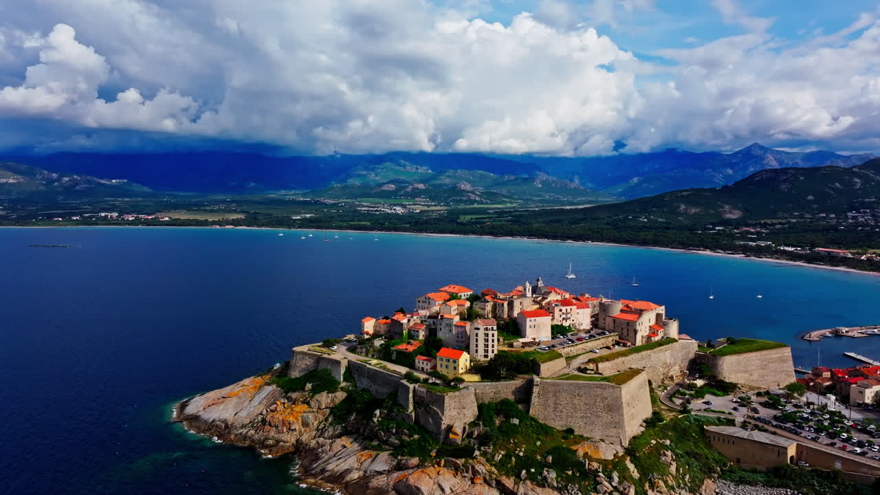 Aerial drone shot over the historic old town of Calvi, Corsica, France. Picturesque view of coastal town, landscape, fluffy clouds, and mountains in the background