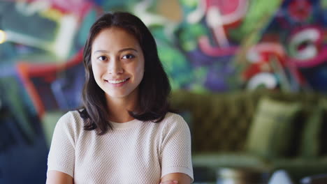 young female asian creative smiling to camera in front of mural in workplace, head and shoulders