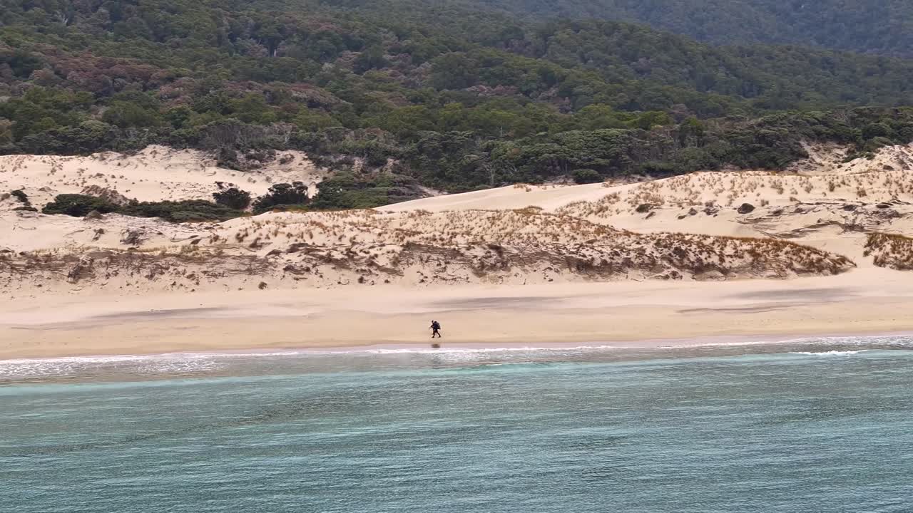 To utforskere vandrer langs en avsidesliggende sandstrand med sanddyner, Stewart Island, Rakiura, tatt med drone.