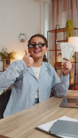 young woman freelancer putting on party glasses holding fan of cash sitting at home office table