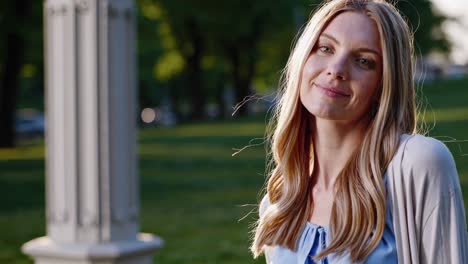 blonde woman wearing light blue shirt and white cardigan standing in sunny park, radiating cheerful expression and elegant style during bright summer day