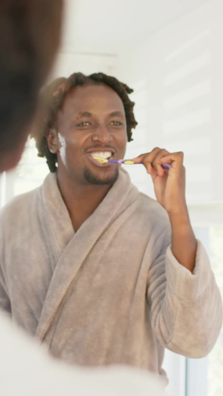 Vertical video: Starting grooming in sunlight, man brushing teeth at mirror in robe with toothbrush