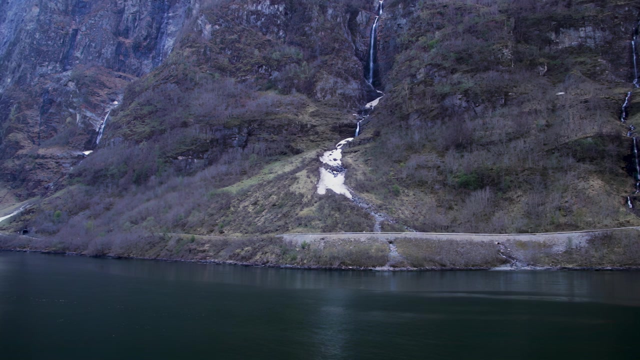 Stejle fjordklipper med smalle vandfald og sneklatter over roligt vand i Norge