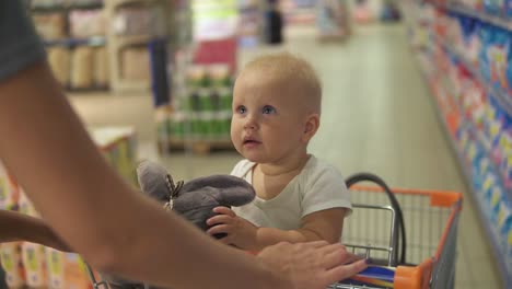 Little-baby-sitting-in-a-grocery-cart-in-a-supermarket-holding-a-toy,-while-her-mother-is-pushing-the-cart-walking-among