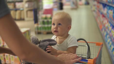 pequeño bebé sentado en un carrito de comestibles en un supermercado con un juguete, mientras que su madre está empujando el carrito caminando entre