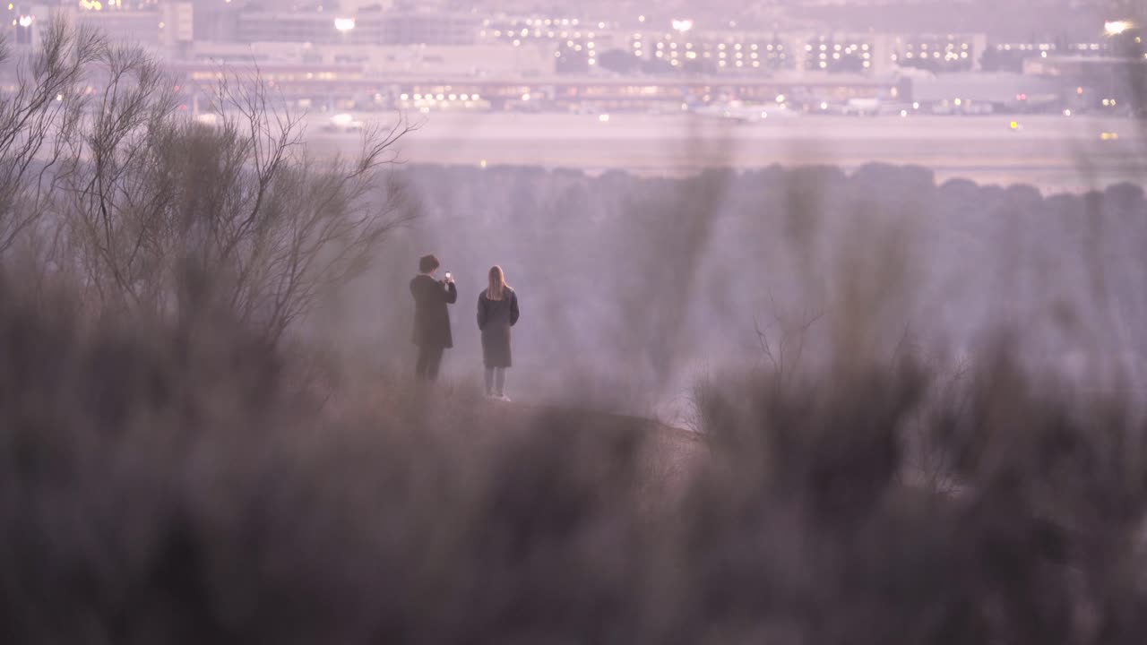 공항과 도시가 있는 언덕 꼭대기에서 스마트폰으로 사진을 찍는 커플의 뒷모습
