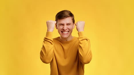 millennial man shaking fists celebrating success posing over yellow background