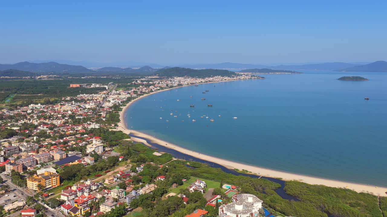 raia da Cachoeira do Bom Jesus di munisipalitas Florianopolis negara bagian Santa Catarina wilayah Selatan Brasil dengan garis pantai teluk melengkung, jalur pesisir perkotaan, dan perahu-perahu yang berlabuh, luncuran drone ke depan