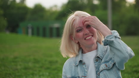 beautiful smiley woman with white bob hair seated on grass laughing hard while watching blurred people playing in park, denim jacket, summer evening light casting warm glow, carefree social moment
