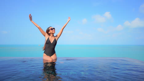 Pretty-young-lady-sitting-on-the-edge-of-infinity-pool-rising-her-arms-above-her-head-with-clear-blue-sky-and-the-ocean-behind-her