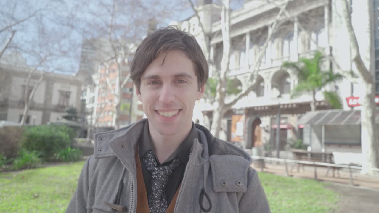 Handsome young house seller man offers keys on the street and smile at camara. Sunny Day outside, park at noon. 4K Medium shot