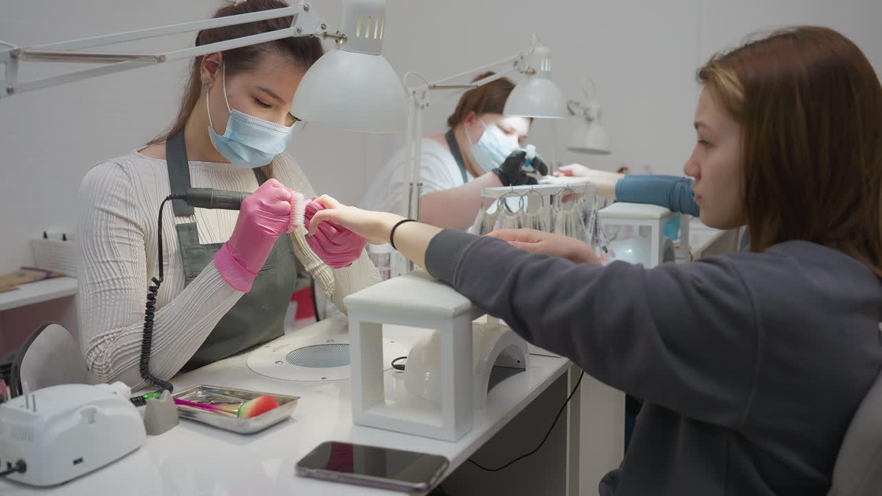 Two nail technicians wearing masks and gloves focused on customer nails under white salon lamps, with customers seated calmly including one with blond hair, manicure tools visible on workstations