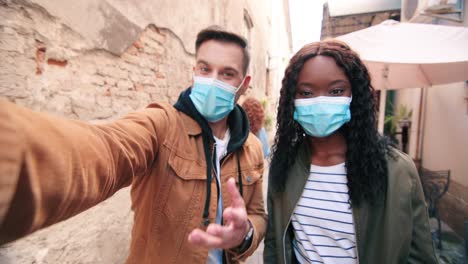 african american woman and caucasian man wearing masks and waving at camera while smiling in the street