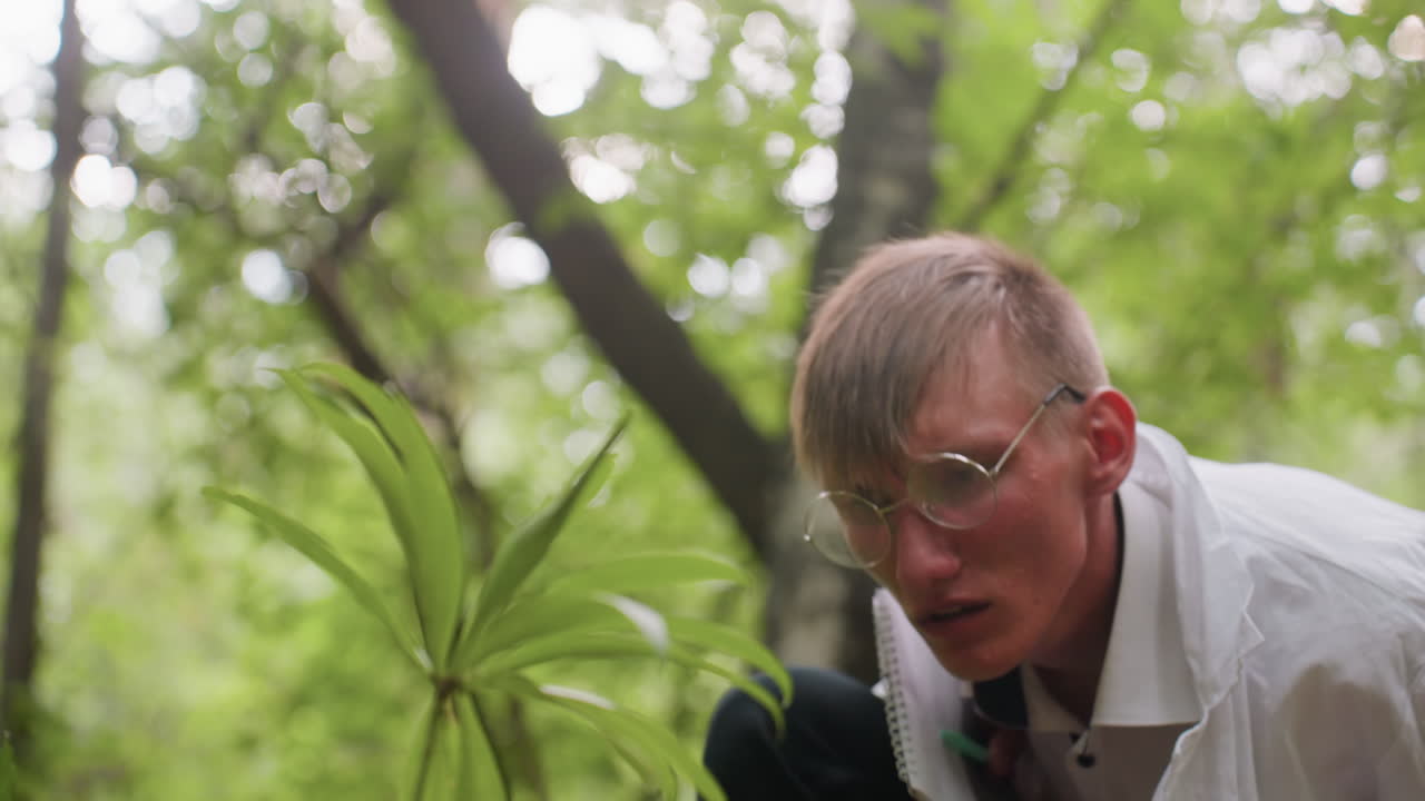 Blurred view of young ecologist in white coat holding jotter bending down to observe flower in forest, focusing on plant study using microscope, ecological research, and environmental documentation