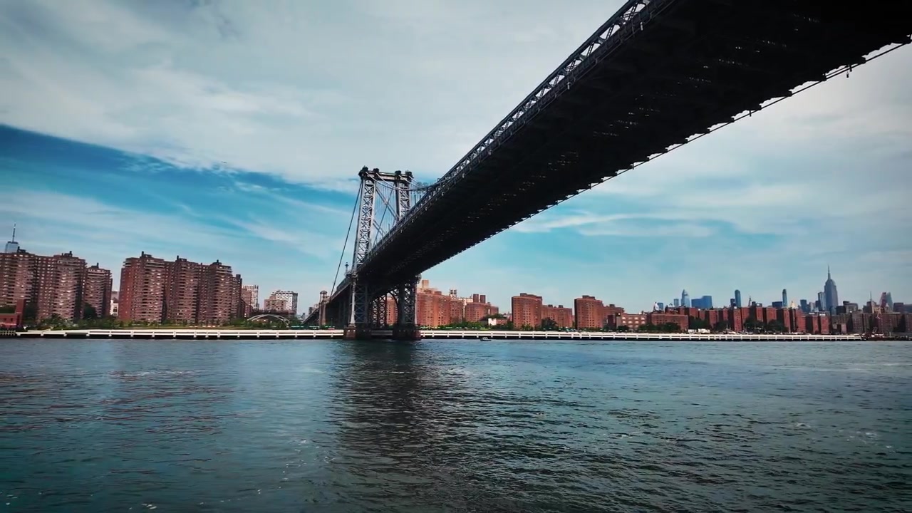 Viaggio in barca cinematografico che si avvicina e passa sotto il Ponte di Manhattan con lo skyline di Lower Manhattan, il One World Trade Center e il lungofiume dell'East River visibili in lontananza.