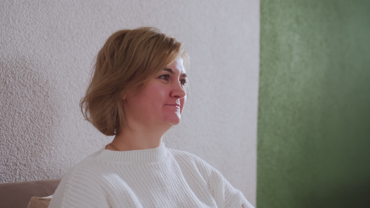 Gentle guest smiles warmly during therapy session on sofa engaging with counselor in cozy interior with soft curtains table evening light creating calm atmosphere highlighting empathic exchange