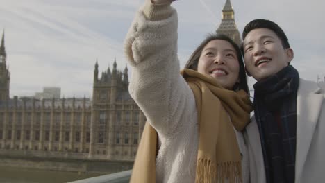 Young-Asian-Couple-On-Holiday-Posing-For-Selfie-In-Front-Of-Houses-Of-Parliament-In-London-UK-1