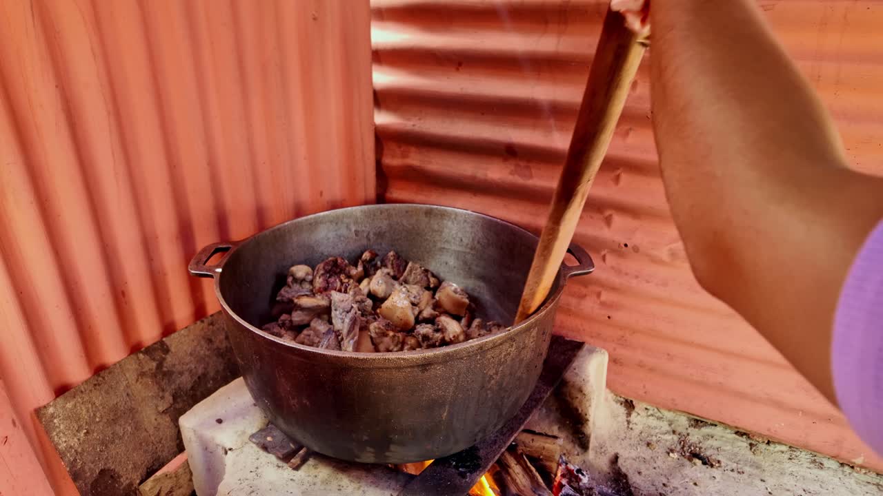 Close-up of a rural Dominican woman frying pork in a metal pot over a wood fire stove in the countryside. The sizzling food is heard under the bright sunlight