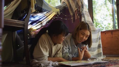 Asian-mother-reading-a-book-for-daughter-under-blanket-fort-at-home