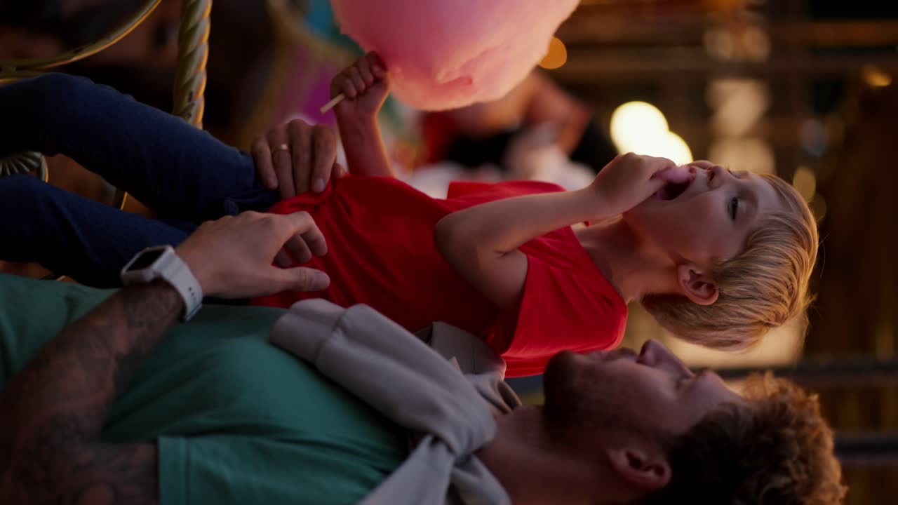 Vertical video of a father and his little blond son with blue hair in a red T-shirt eating pink cotton candy in Luna Park in the evening