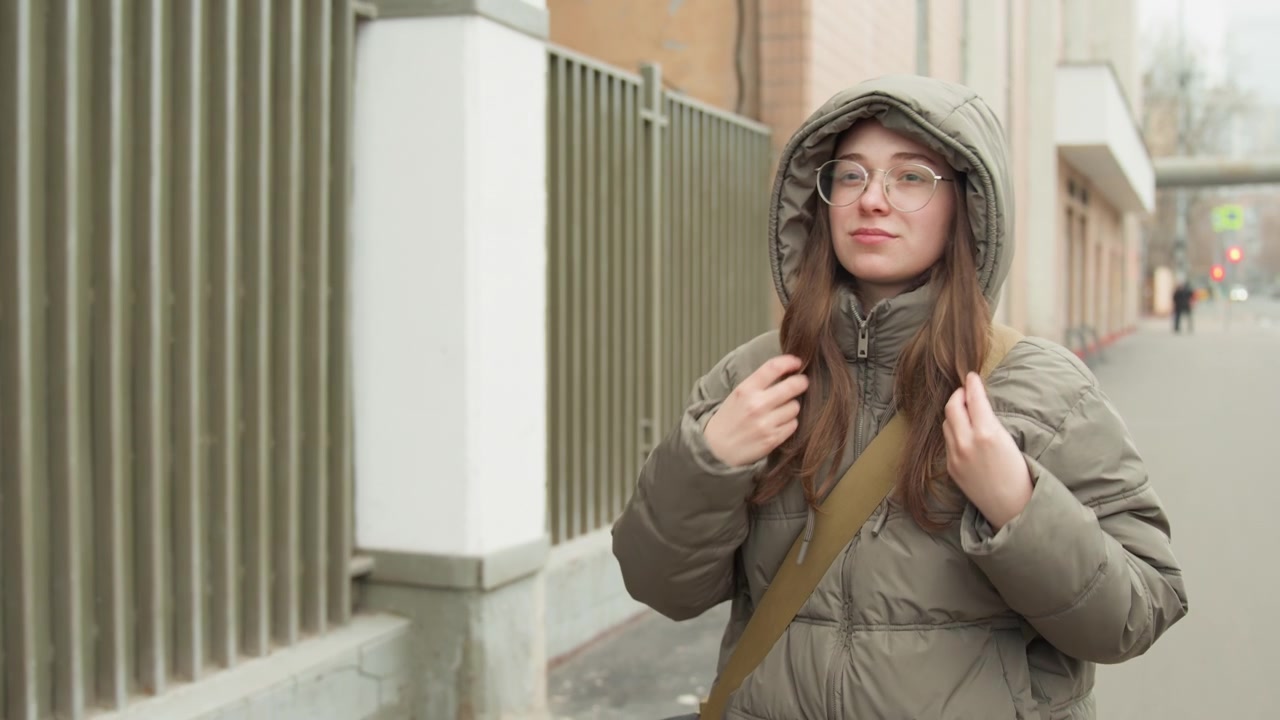 Young woman adjusting hood on sidewalk, green puffer coat, clear glasses and shoulder bag, hands tucking hair beside metal fence, casual upbeat morning vibe with soft overcast light and candid smile