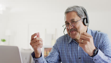 video of happy senior biracial man sitting at desk using headset and laptop making video call