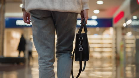 low angle lower view of shopper carrying designer bag strolling down glossy tiled mall hall under bright lights in modern retail corridor wearing jeans and chunky black boots capturing dynamic motion