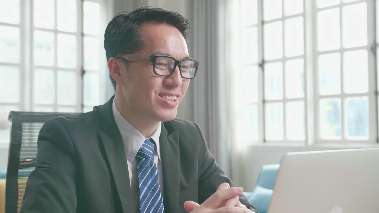 un hombre de negocios asiático con gafas y traje de negocios haciendo una llamada de video en la computadora mientras trabaja en casa.