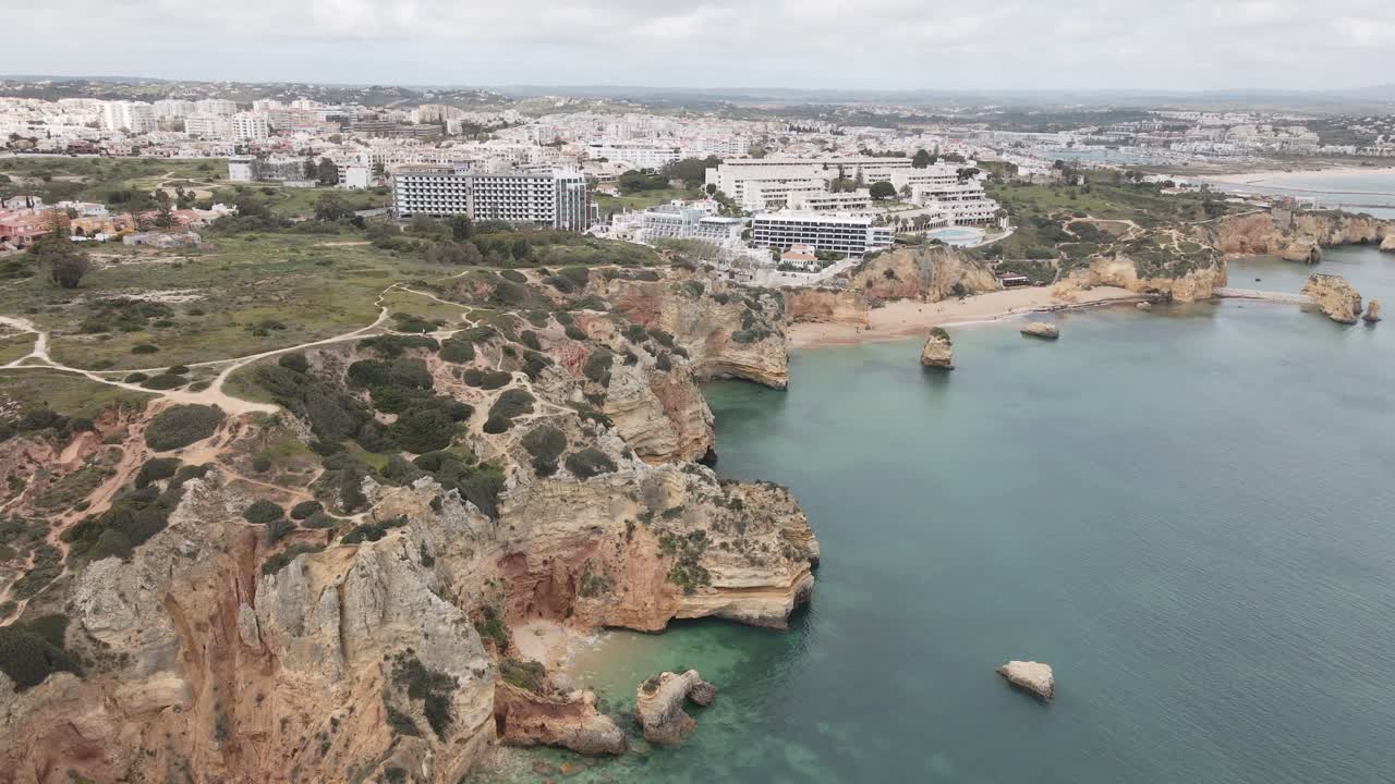 Aerial drone ascends, revealing turquoise sea, dramatic cliffs, and Lagos city skyline in Algarve
