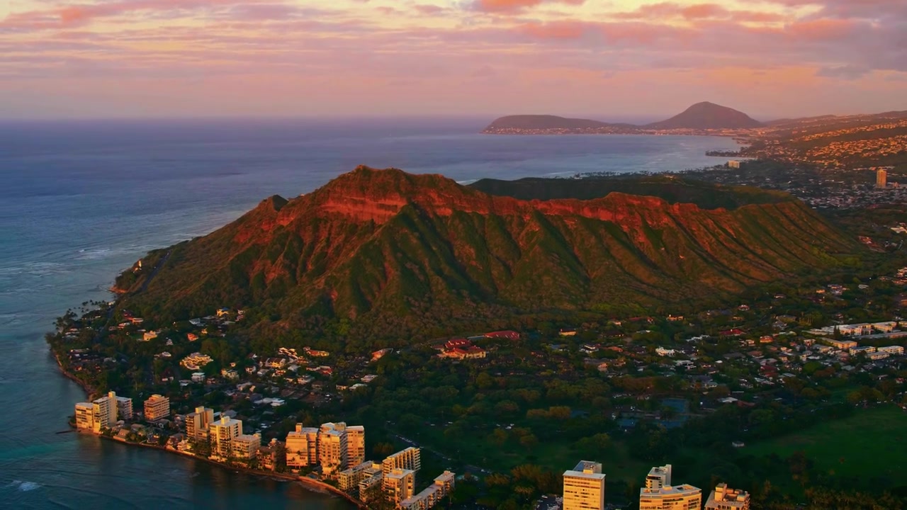 Una prospettiva aerea cinematografica mozzafiato all'ora d'oro che scivola con grazia sopra l'iconico cratere vulcanico Diamond Head, affacciandosi sulla scintillante costa e sullo skyline di Waikiki, Hawaii.