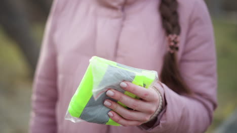 close-up of woman in pink quilted winter jacket holding folded neon yellow and gray reflective safety vest in transparent plastic bag outdoors, symbolizing preparedness, protection, in outdoor activities