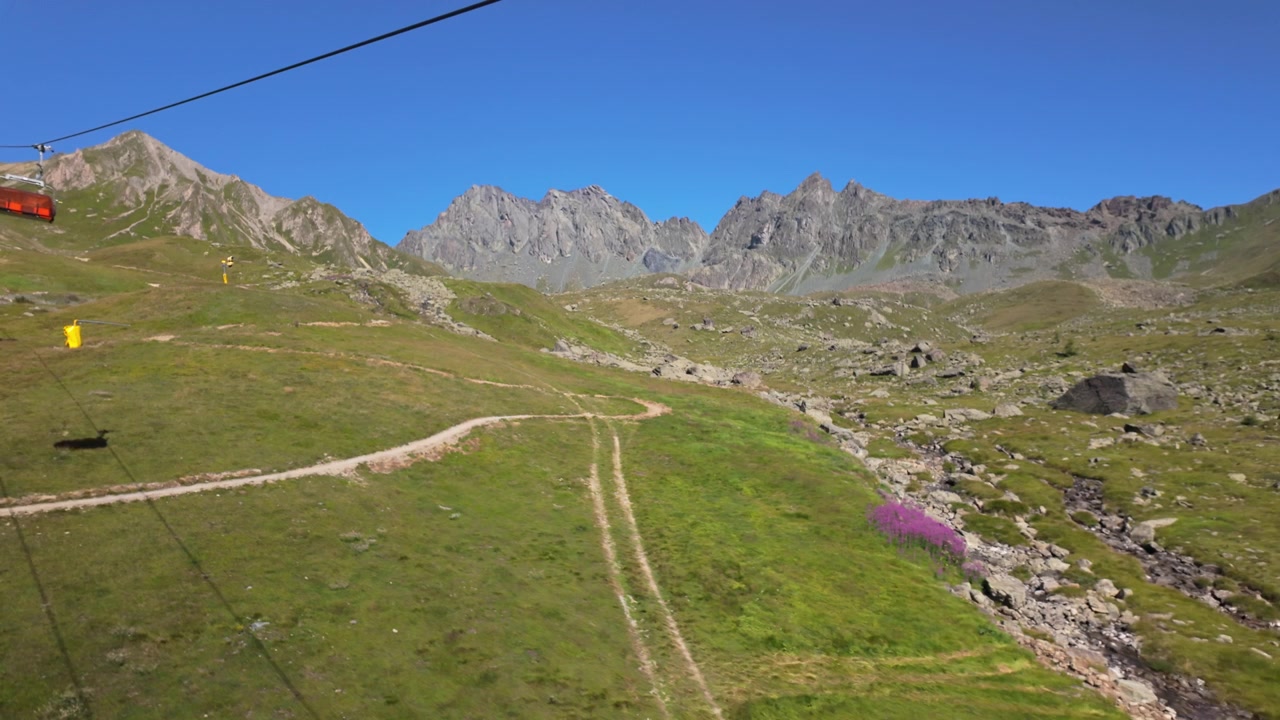 Cresta montuosa rocciosa sopra verdi prati alpini, vista da una funivia con un sentiero tortuoso, un piccolo ruscello e un cielo estivo limpido e blu
