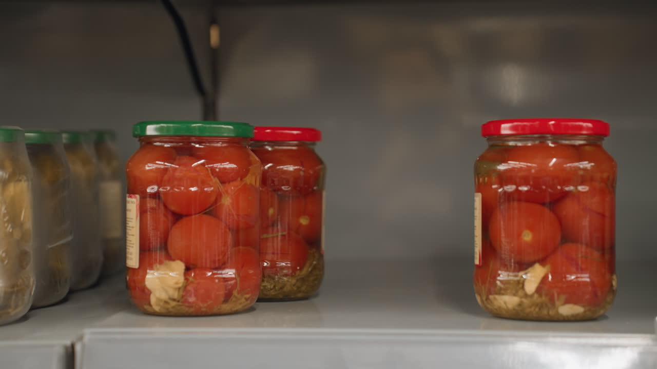 Close up of shopper hand grabbing transparent glass jar of tomatoes with green lid from metal shelf rails in bright grocery aisle highlighting blurred movement and polished nails wrist watch