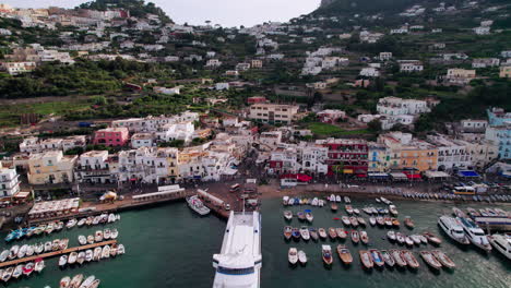 A-cinematic-wide-drone-shot-revealing-Marina-Grande-on-the-island-of-Capri-in-Campania,-Italy,-showing-a-ferry-and-boats-in-the-port,-tourists-strolling-the-coast,-and-colorful-pastel-buildings