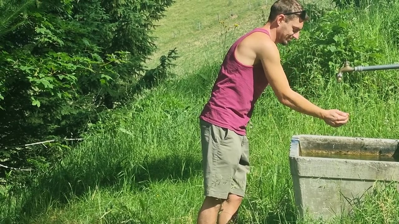 A man cools off by washing his arms in a cattle drinking fountain amid the lush summer scenery of the Swiss Alps, capturing alpine heat, fresh mountain water, outdoor recreation and rural life