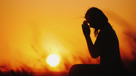 silhouette of a woman praying against the background of an orange sky and a large setting sun