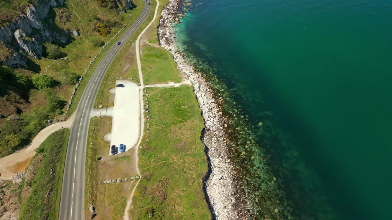 Wide overhead aerial video of the Causeway Coastal Route in County Antrim, Northern Ireland, UK, on a bright and sunny day. A car passes below. Filmed in 4K, 60FPS and with Rec709 color.