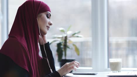 Gorgeous-arabic-looking-female-with-hijab-puts-headphones-in-her-ears-and-starts-listening-to-her-favourite-music.-Indoors-footage