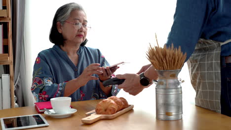 active senior asian woman working in a coffeeshop using a mobile device, smartwatch for nfc, qr code scanning, contactless payment to pay for coffee and snack