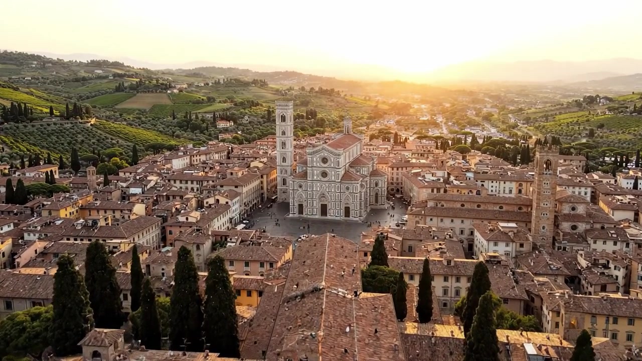 Veduta aerea di una piazza storica al tramonto, circondata da dolci colline e vegetazione lussureggiante, che mostra un'architettura mozzafiato e strade affascinanti