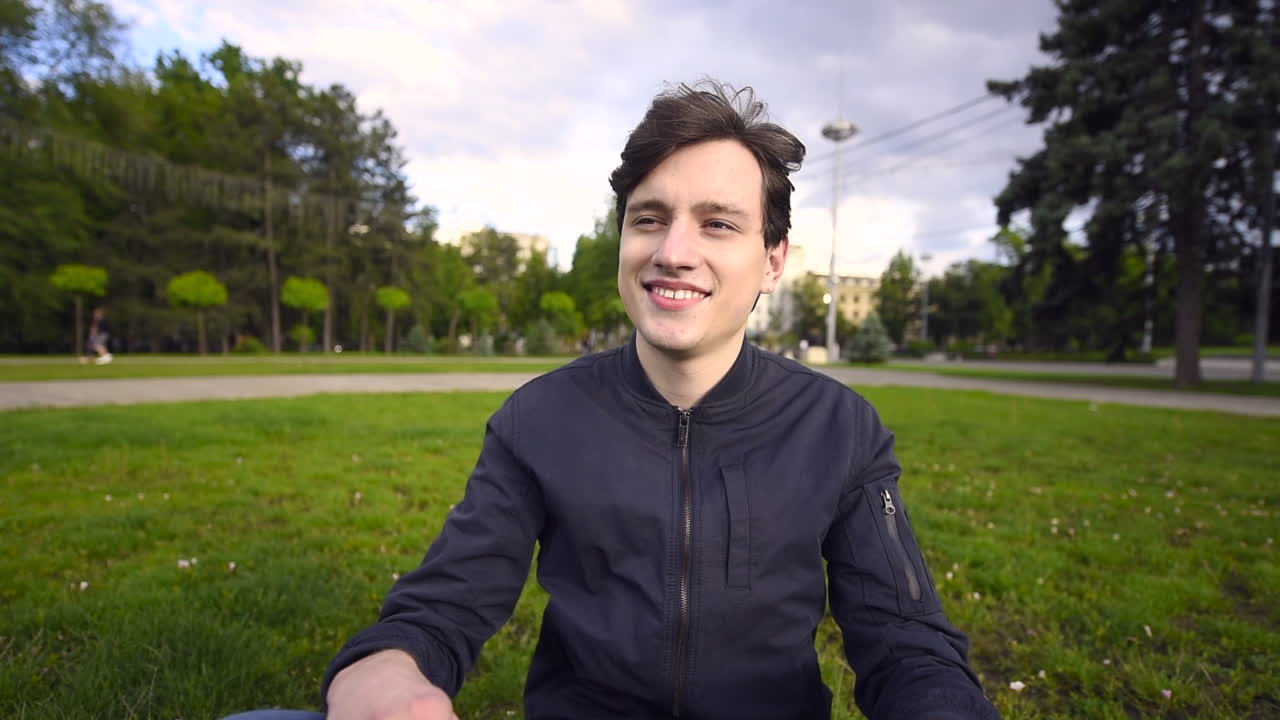Satisfied young man removing a black protective mask from his face and looking around while resting on the grass in park. Driving cars on background at sunset. Quarantine and Corona Virus idea