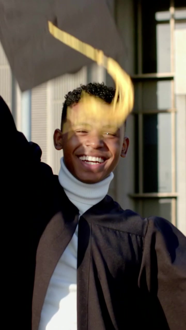 Vertical video: Smiling graduate raising arm after cap toss outside campus, wearing black gown