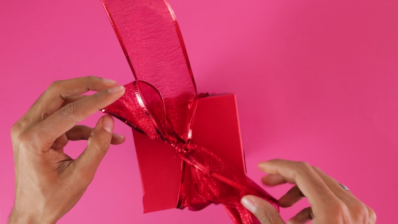 Tying red ribbon bow around gift box for Valentine, pink background
