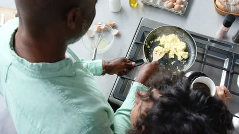 happy biracial couple cooking and preparing scrambled eggs on pan in kitchen, slow motion
