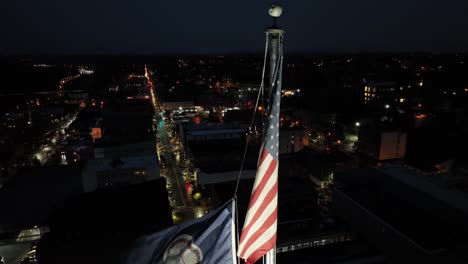 aerial close up of american flag and flag of virginia state on flagpole. illuminated main street of american town at night. cityscape with buildings and homes in background. proud state of usa.