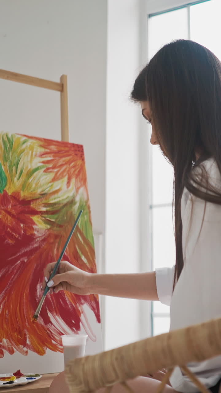Thoughtful woman artist at work upon impressive picture of colorful exotic flowers with red petals on canvas in rattan armchair in studio backside view