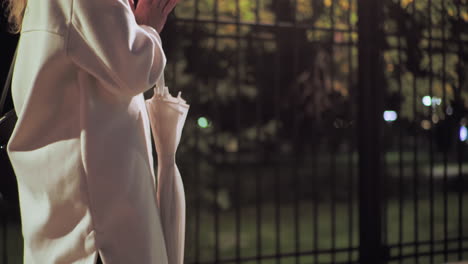 young girl strolling beside iron fence under soft streetlight, gazing thoughtfully to right, gentle light highlighting curly hair, bokeh lights and blurred greenery