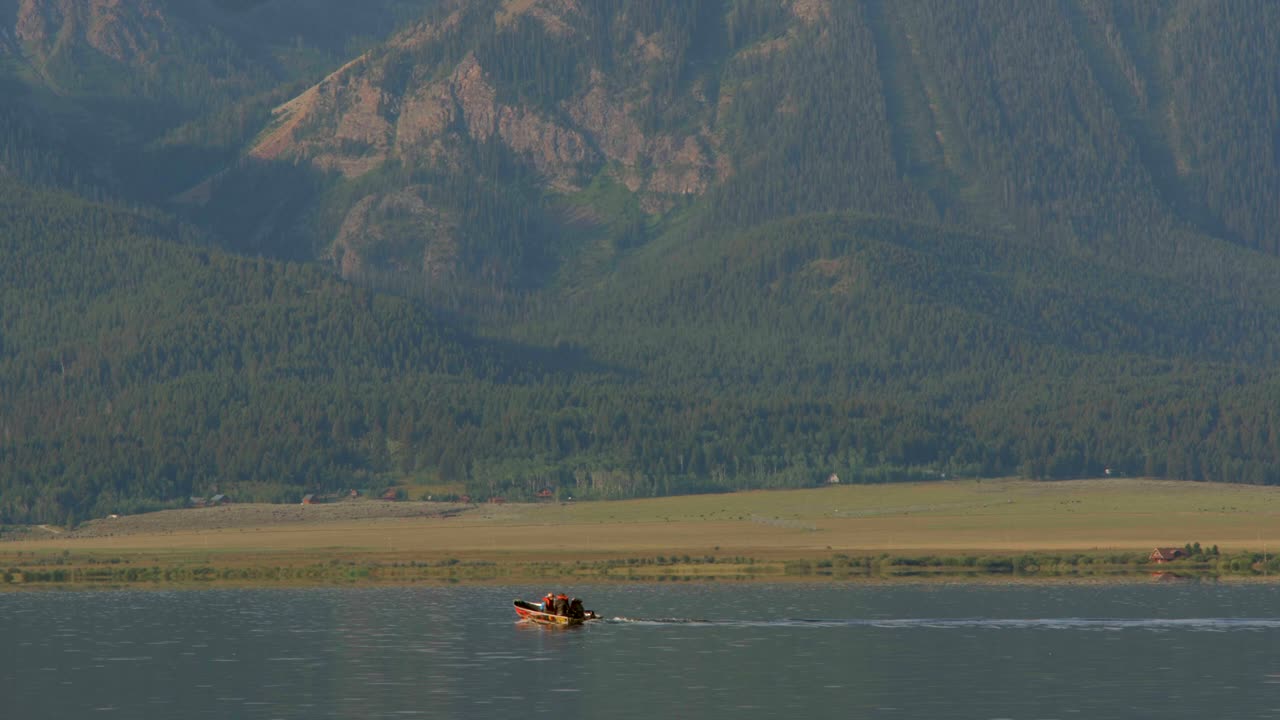 A raft is seen floating from a distance on a river. A large mountain is in view covered with pine trees.